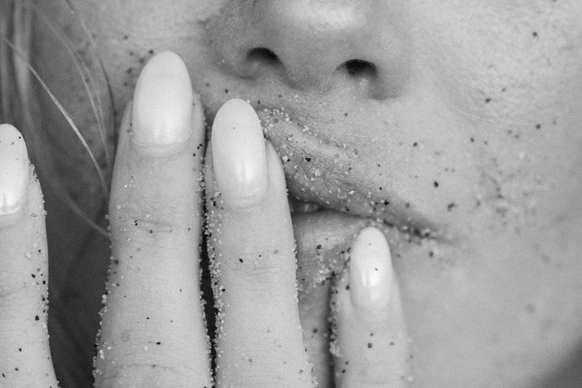 A close-up black and white photograph of a woman's lower face and hand. Her hand, with neatly manicured nails, is pressed against her lips, and both her hand and lips are sprinkled with fine grains of sand